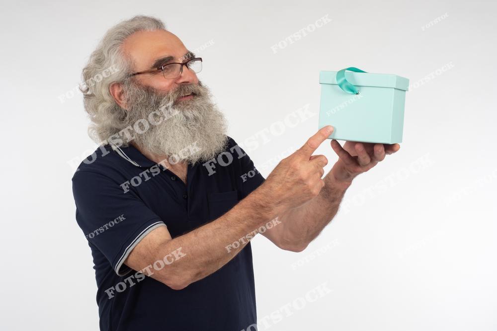 На фотографии изображен пожилой мужчина с густой бородой, держащий в руках голубую подарочную коробку и указывающий на нее пальцем. Мужчина выглядит радостным и довольным, демонстрируя коробку с подарком.